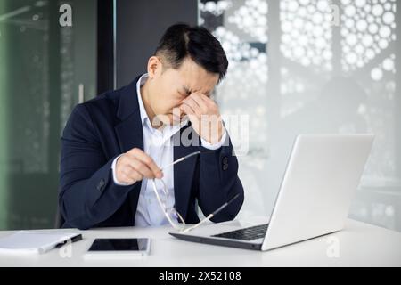 Un homme d'affaires asiatique d'âge moyen se sent accablé et stressé dans un cadre de bureau moderne, massant le pont de son nez tout en regardant son ordinateur portable. Banque D'Images