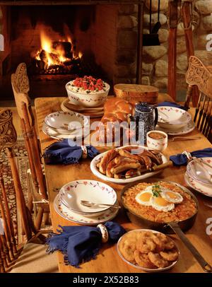 Petit déjeuner composé d'œufs, rosti, saucisses grillées, beignets de pommes et pain maison sur une table en pin rustique devant une cheminée en pierre. Banque D'Images