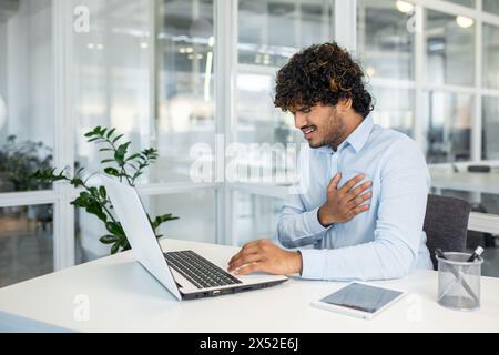 Un homme dans un bureau moderne ressent une douleur thoracique soudaine en utilisant son ordinateur portable, mettant en évidence des problèmes de santé potentiels liés au stress lié au travail. Banque D'Images