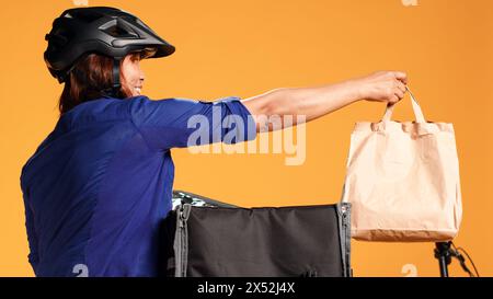 Gros plan d'un transporteur asiatique proposant aux clients de commander des sacs à lunch. Travailleur amical de livraison de nourriture arrivant rapidement à l'adresse du client, prenant le repas hors du sac à dos thermique, isolé sur fond de studio Banque D'Images