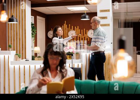 Homme caucasien senior debout au comptoir du hall de l'hôtel et s'enregistre avec passeport. Concierge femme au bureau d'enregistrement retournant le document personnel du voyageur âgé après vérification de l'identité. Banque D'Images