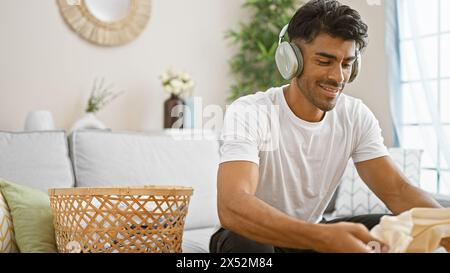 Homme hispanique pliant le linge à la maison tout en écoutant de la musique sur son casque capture un moment domestique détendu. Banque D'Images