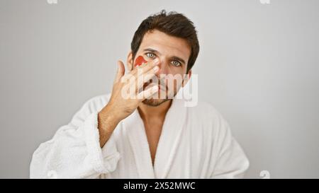 Jeune homme hispanique en robe blanche applique la crème de soin du visage sur fond blanc isolé Banque D'Images