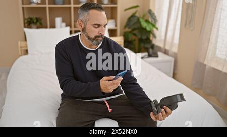 Homme barbu mature dans la chambre à coucher à l'aide d'un smartphone et tenant des écouteurs, représentant la technologie et le style de vie. Banque D'Images
