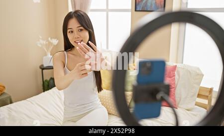 Une jeune femme asiatique enregistre un vlog avec un smartphone dans une chambre confortable, en utilisant une lumière annulaire pendant qu'elle fait des gestes. Banque D'Images