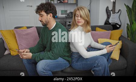 Une femme jette un regard sournois sur le téléphone d'un homme tandis que les deux s'assoient dos à dos sur un canapé à l'intérieur avec des oreillers colorés. Banque D'Images