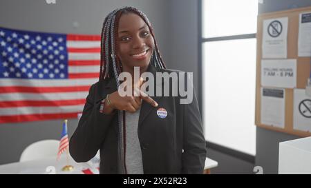 Une femme afro-américaine souriante avec des tresses montre son autocollant «j'ai voté» dans une pièce avec un drapeau américain. Banque D'Images