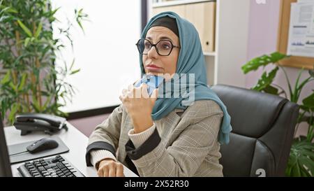 Une femme d’âge moyen en hijab écoute attentivement sur un smartphone dans son bureau moderne, entourée de plantes et de technologies d’intérieur. Banque D'Images