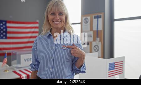 Une femme blonde souriante dans une chemise rayée montre son autocollant «j'ai voté» dans un bureau de vote américain avec des drapeaux américains. Banque D'Images