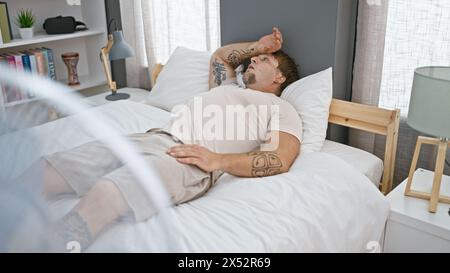 Un jeune homme barbu se prélassant dans son lit, avec des tatouages visibles, à l'intérieur d'une chambre moderne faisant subtilement allusion à la détente et au confort. Banque D'Images