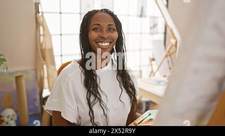 Portrait d'une jeune femme souriante aux cheveux bouclés, habillée artistiquement, dans un cadre de studio d'art ensoleillé. Banque D'Images