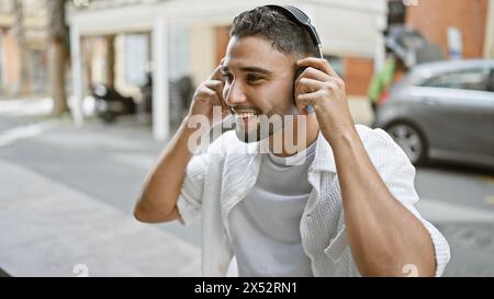 Homme barbu souriant portant des écouteurs dans un cadre urbain ajuste sa musique par une journée ensoleillée. Banque D'Images