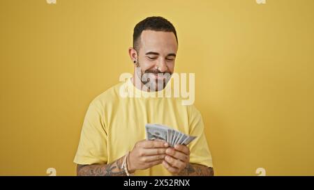 Beau jeune homme hispanique avec une barbe sourit alors qu'il compte l'argent sur un fond jaune isolé. Banque D'Images