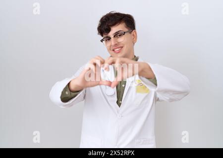 Jeune homme non binaire portant l'uniforme de médecin et stéthoscope souriant dans l'amour faisant la forme de symbole de coeur avec les mains. concept romantique. Banque D'Images