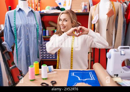 Jeune femme caucasienne couturière designer travaillant à l'atelier souriant dans l'amour faisant la forme de symbole de coeur avec les mains. concept romantique. Banque D'Images