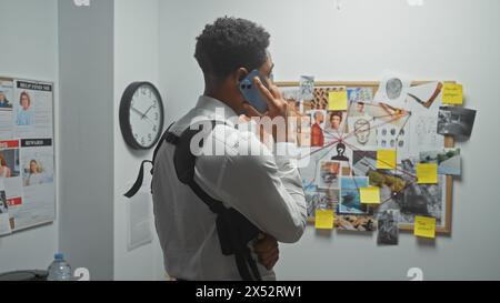 Un homme dans le bureau d'un détective parle au téléphone à côté d'un panneau de preuves avec des photos et des notes. Banque D'Images