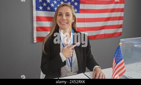 Une jeune femme caucasienne souriante montre son autocollant «j'ai voté» dans un centre de vote intérieur américain avec des drapeaux. Banque D'Images