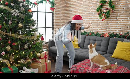 Jeune femme hispanique avec chien chihuahua jouant et dansant portant un chapeau de noël à la maison Banque D'Images