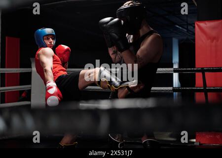 Jeune boxeur en sportswear noir défendant de son rival donnant bas coup de pied par jambe dans le shinguard protecteur pendant le combat de sparring sur le ring de boxe Banque D'Images