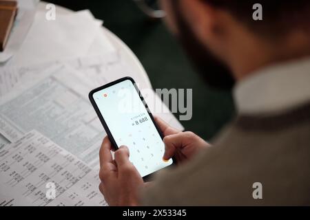 Smartphone dans les mains du jeune homme avec le résultat de calculs à l'écran sur les factures financières et autres documents couchés sur la table Banque D'Images