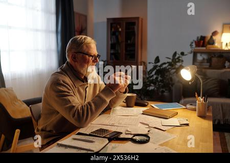 Vue latérale de l'homme barbu senior dans des lunettes à l'aide de la calculatrice dans le smartphone tout en étant assis à la table avec des factures de paiement de logement Banque D'Images