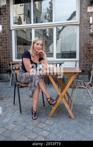 Cette image représente une femme blonde sereine assise seule à une table de café en bois sur un trottoir pavé. Elle se penche doucement sur son coude, son menton reposant sur sa main, dépeignant une expression pensive. La femme est vêtue d'un haut noir élégant et sans manches associé à une jupe à rayures zébrées, complétée par des sandales noires à lanières. Derrière elle, la grande fenêtre du café reflète un vieux bâtiment en pierre, ajoutant une ambiance historique au cadre urbain. La lumière de fin d'après-midi suggère une fin paisible à une journée animée. Femme contemplative assise à une table de café extérieure. Photo de haute qualité Banque D'Images