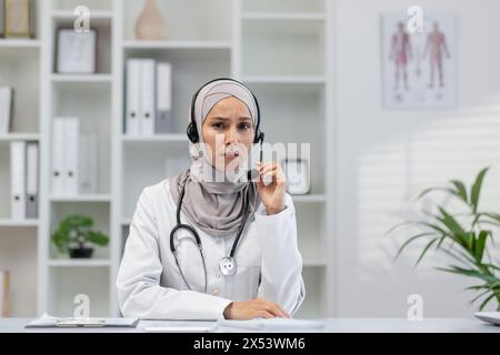 Une femme médecin en hijab est assise à son bureau, paraissant inquiète tout en tenant un casque. Elle semble écouter attentivement, donnant probablement une consultation. Banque D'Images