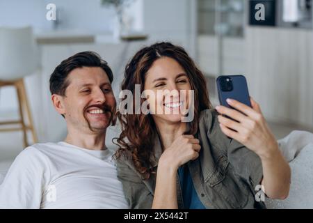 Couple joyeux prenant selfie avec smartphone, dans un espace de vie moderne lumineux et aéré. L'homme fait des moustaches drôles avec les cheveux de la femme. Banque D'Images