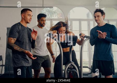 Jeunes adultes multiraciaux en vêtements de sport applaudissant pour le vélo d'ami sur le vélo d'exercice à la salle de gym Banque D'Images