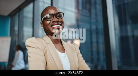 Femme d'affaires dans une réunion de conseil. Elle a l'air heureuse et réussie, portant des lunettes et debout dans un bureau panoramique. Son sourire professionnel respire Banque D'Images