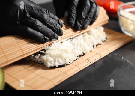 Chef en gants enveloppant le rouleau de sushi à la table texturée sombre, gros plan Banque D'Images