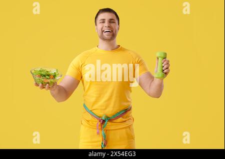 Jeune homme avec des rubans à mesurer, salade et haltère sur fond jaune. Concept de perte de poids Banque D'Images