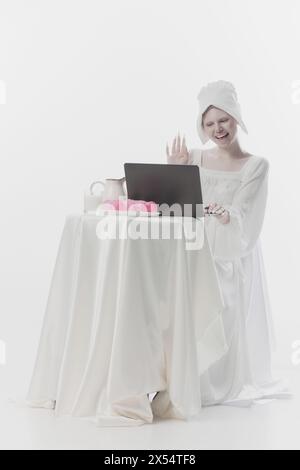 Jeune femme attrayante habillée comme une simple femme paysanne agitant bonjour tout en se réunissant en ligne à l'ordinateur portable sur fond blanc de studio. Banque D'Images