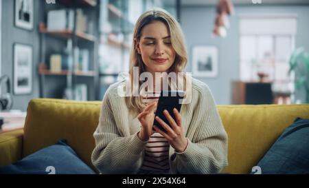 Belle femme caucasienne utilisant smartphone dans le salon élégant tout en se reposant sur un canapé confortable. Jeune femme à la maison, naviguer sur Internet, utiliser les réseaux sociaux, s'amuser à plat. Banque D'Images