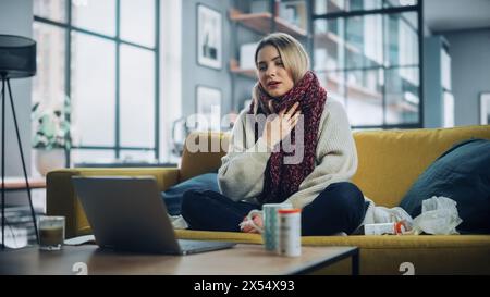 Jeune femme malade se soufflant le nez et appelant au bureau avec appel téléphonique vidéo de Cozy Living Room. Femme malade et nauséeuse dans l'écharpe chaude parlant à un ami sur une caméra Internet en direct. Banque D'Images