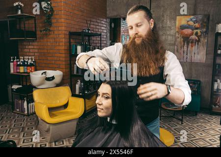 Coiffeur peignes une femme. Homme jeune coiffeur positif enthousiaste avec une longue barbe et les cheveux en gilet, peignant soigneusement la femme cliente dans le fauteuil i. Banque D'Images