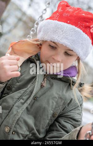 Sympathique jeune jolie fille sur une aire de jeux avec un chapeau de Père Noël rouge chic. En hiver, juste avant Noël, les cadeaux suscitent une grande excitation. Banque D'Images