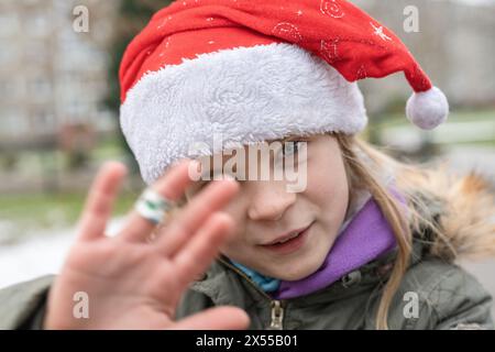 Sympathique jeune jolie fille sur une aire de jeux avec un chapeau de Père Noël rouge chic. En hiver, juste avant Noël, les cadeaux suscitent une grande excitation. Banque D'Images