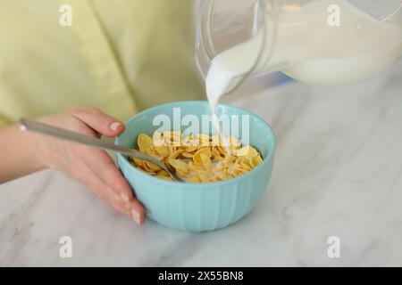 Préparer le petit déjeuner. Femme versant le lait de la cruche dans le bol avec des flocons de maïs à la table en marbre blanc, gros plan Banque D'Images