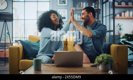 Couple multiethnique diversifié sont assis sur un canapé canapé dans le salon élégant et choisir des articles à acheter en ligne avec ordinateur portable, Give High Five. Des amis ou des collègues discutent de projets de travail. Banque D'Images