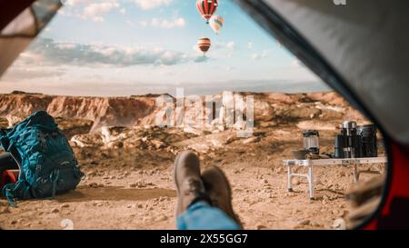 POV d'un touriste se reposant dans une tente en plein air au sommet d'une montagne rocheuse avec Canyon Valley et volants montgolfières dans le ciel. Backpacker portant des bottes de randonnée et un Jean Dusty Traveller. Banque D'Images