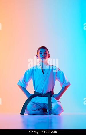 Adolescent, athlète de karaté en uniforme, kimono blanc et ceinture verte assis sur fond bleu orange dégradé dans la lumière néon Banque D'Images