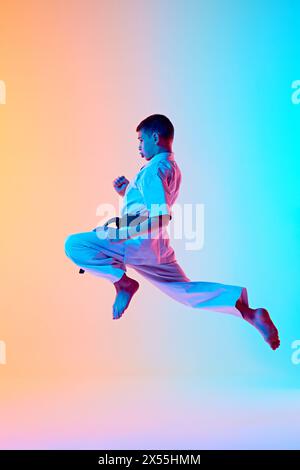 Jeune athlète engagé dans l'entraînement de karaté, sautant dans la pose dynamique sur fond bleu orange dégradé dans la lumière néon Banque D'Images