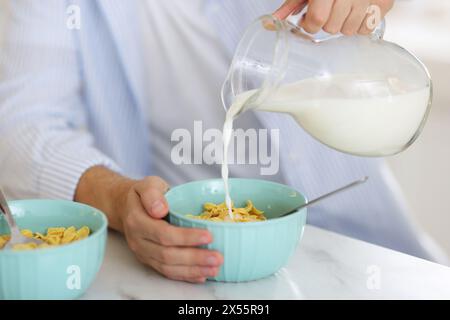 Préparer le petit déjeuner. Homme versant le lait de la cruche dans le bol avec des flocons de maïs à la table en marbre blanc, gros plan Banque D'Images