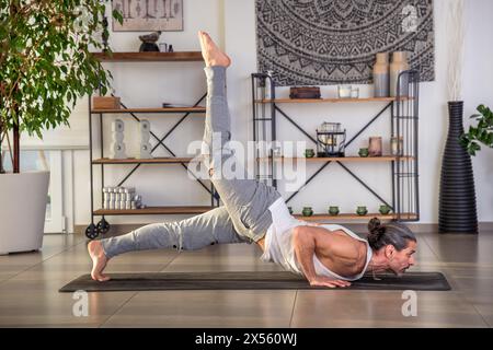 Vue latérale d'un homme adulte en vêtements de sport regardant loin tout en étant allongé sur le tapis dans la pose Ardha Shalabhasana et faire du yoga dans un hall spacieux avec des fournitures d'art. Banque D'Images