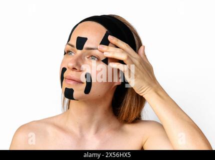 Rajeunissement facial et thérapie alternative anti-âge. Modèle de beauté femme Brunette appliquant des bandes faciales kinesio sur son visage devant le dos blanc Banque D'Images