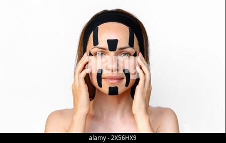 Rajeunissement facial et thérapie alternative anti-âge. Modèle de beauté femme brune isolé appliquant des bandes faciales kinesio sur son visage devant whi Banque D'Images