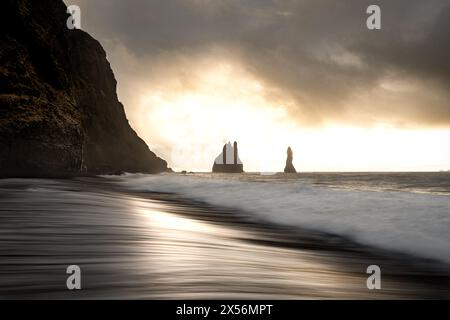 Lever de soleil doré derrière Hálsanefshellir empilements de mer, vagues peignant des stries lumineuses sur la plage de sable noir. Capture longue exposition de la beauté de l'aube. Banque D'Images