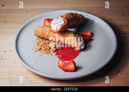 Un cannoli rempli de crème, accompagné de fraises et d'une garniture émiettée, servi sur une élégante assiette grise. Banque D'Images