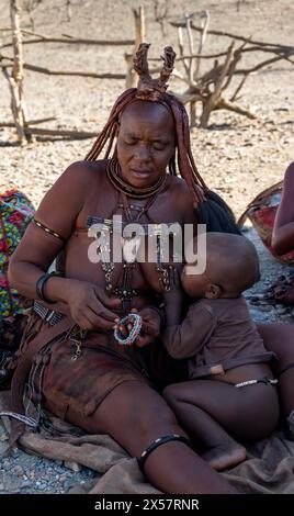 Femme Himba mariée traditionnelle allaitant son bébé, faisant des bracelets, près d'Opuwo, Kaokoveld, Kunene, Namibie Banque D'Images